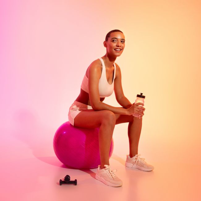 A young, happy woman sitting on top of an exercise ball sipping water and smiling, taking a break from her Reformer Pilates workout at Silverlink Studio 24/7 Reformer Pilates